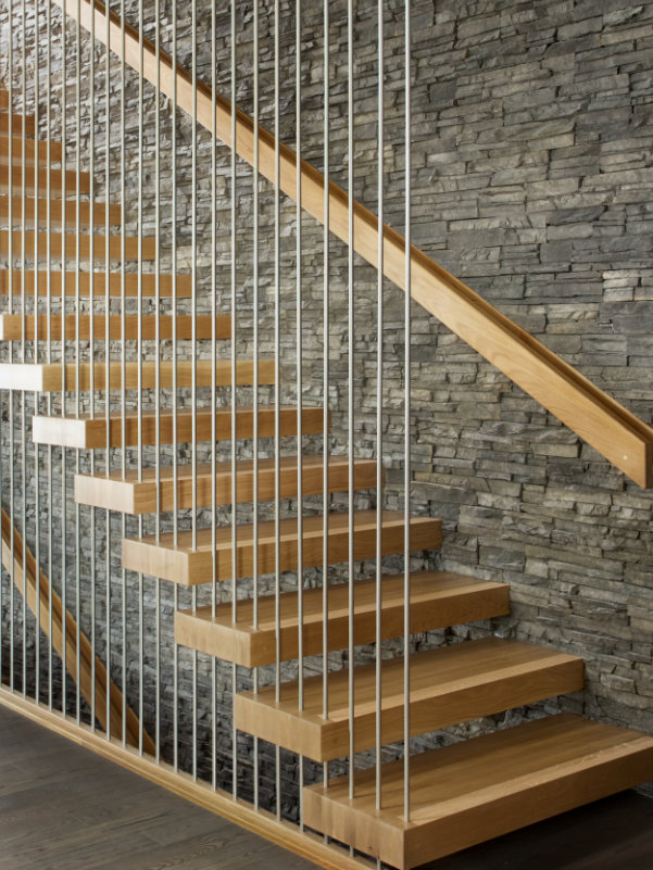A cantilevered white oak staircase against a rock wall with floor to ceiling steel tube railing.